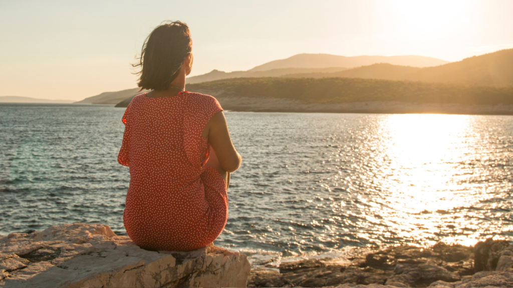 Woman sitting on a rock overlooking water at sunset, reflecting quietly